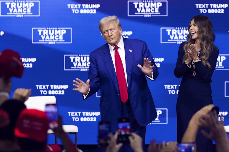 donald trump on stage while kristi noem looks on and applauds