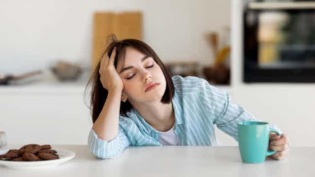 Uma jovem adormecida em uma cozinha, com uma xícara de café na mão.