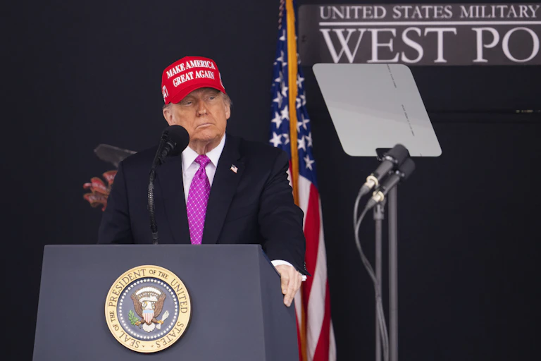 A man in a red hat and suit stands at a lectern with the presidential seal on it.