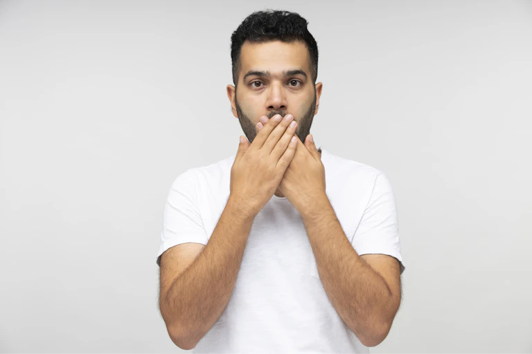 A man in a white t-shirt covering his mouth with both hands.