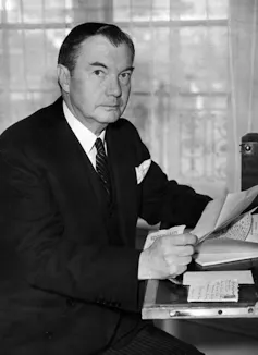 A dark haired man in a suit, sitting at a desk with papers in his hand.