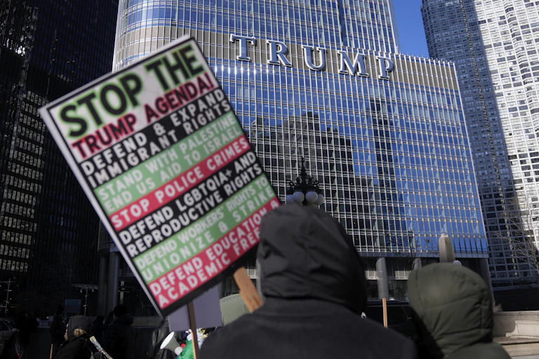 A proteser carries a sign listing grievances against Donald Trump with the Trump Tower in the background.