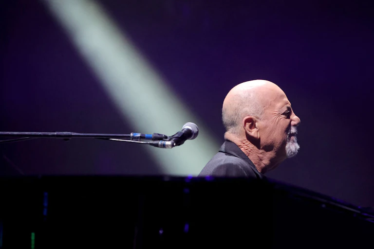 Side profile shot of Billy Joel on stage behind a piano and beside a mic, a beam of light shining obliquely down on his back