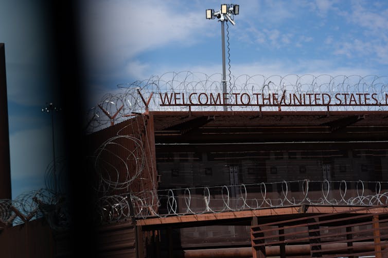 A sign says 'Welcome to the United States' surrounded by barbed wire on top of a building on a clear day.