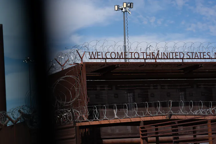 A sign says 'Welcome to the United States' surrounded by barbed wire on top of a building on a clear day.