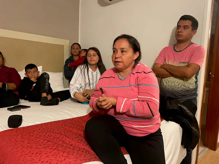 Woman sitting on hotel bed, man standing by the bed and women and several children sitting behind her on it