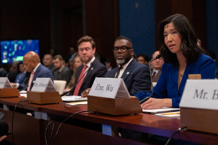 An Asian woman with a placard in front of her is seated and speaks into a microphone, seated next to several men who are also dressed formally and seated in front of microphones.