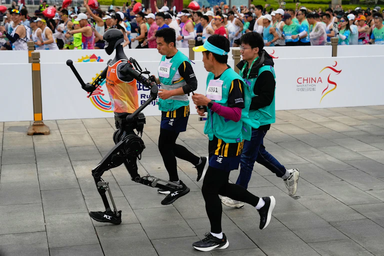 a humanoid robot runs with three people on one side of a fence while a crowd of runners goes by on the other side