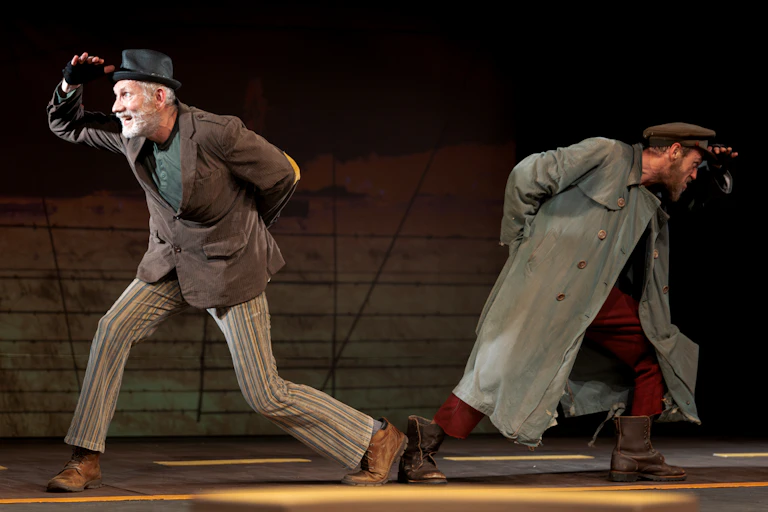 Two bearded men on a stage in hats and scruffy clothes; each holds their hands to their foreheads, looking into the distance.