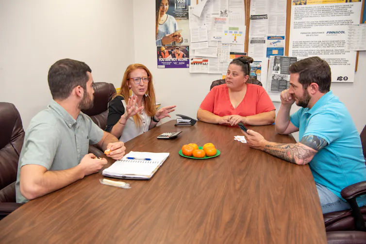Four people sit and talk at the end of a conference table with posters in the background.