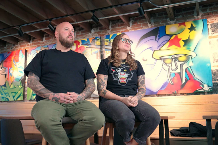 A man and woman sit on stools in front of pop art at their restaurant in Denver.
