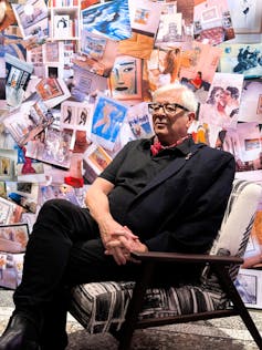El hombre con cabello blanco estĆ” sentado en una silla con una pared trasera cubierta de dibujos.