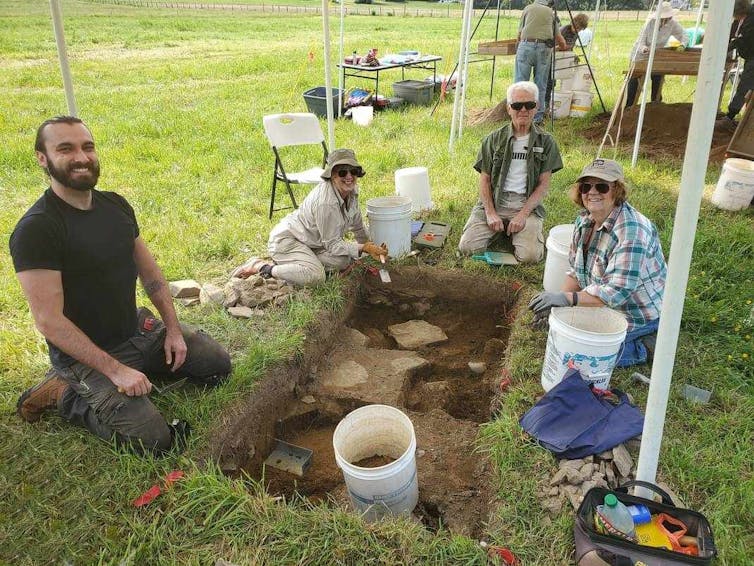 several people seated around rectangular hole showing dirt that's cut into grass