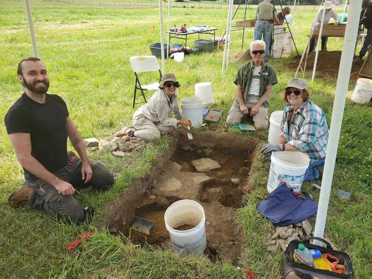 several people seated around rectangular hole showing dirt that's cut into grass