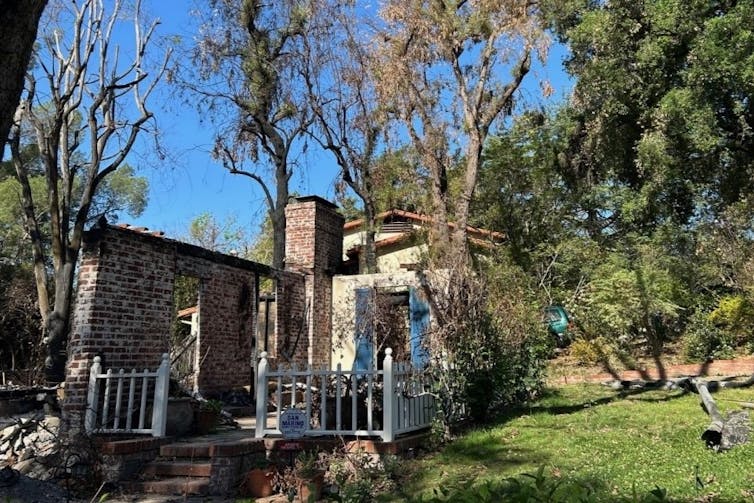 A burned home, with only brick walls standing in some areas, with a green lawn and shrubs in adjacent to the house.