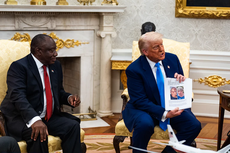 A white older man in a navy blue suit holds up a white paper with two people's faces on it as he sits next to an older Black man wearing a black suit and red tie.