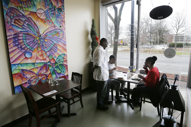 A man in a white chef's coat stands with his hands behind his back as he speaks to two women sitting and eating. There is artwork with purple, orange and pink butterflies and an empty table nearby.
