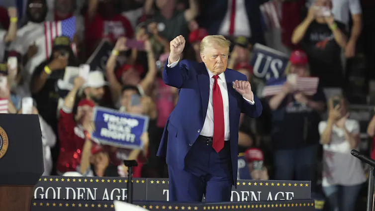An elderly rotund orange-hued white man with a golden comb over wearing a suit and long red tie dances on a stage with a crowd of people behind him.