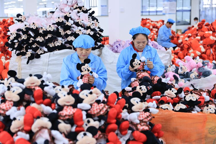 Two female workers wearing blue surrounded by piles of stuffed animals.