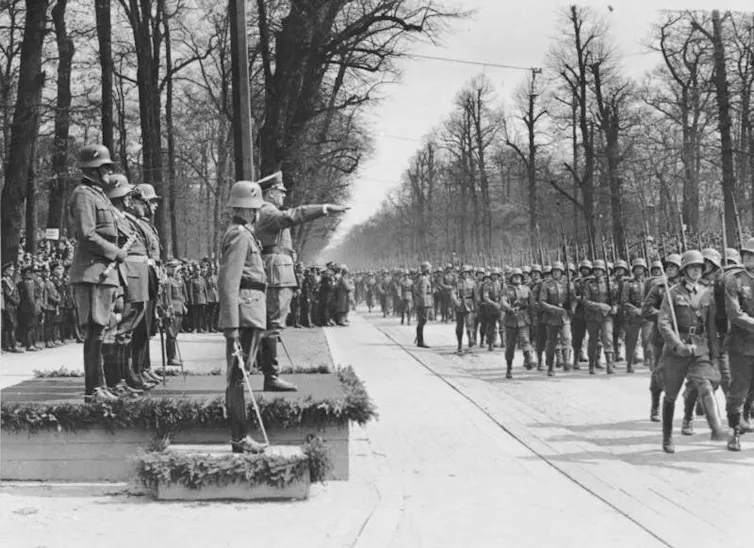 a black and white photo of soldiers marching in a parade. A man on a platform gives a Nazi salute.