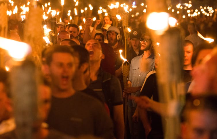 A group of men hold torches and stand together and appear to be shouting, with their mouths open.