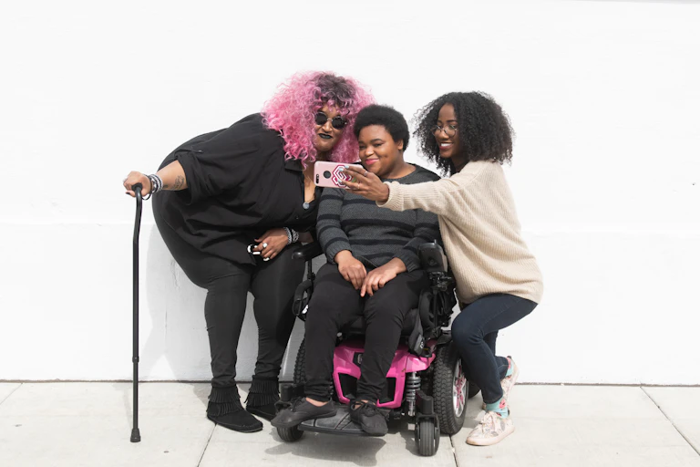 Three Black and disabled friends (a non-binary person with a cane and tangle stim toy, a non-binary person sitting in a power wheelchair, and an invisibly disabled femme) smiling and taking a cell phone selfie together. All are outdoors in front of a white wall.
