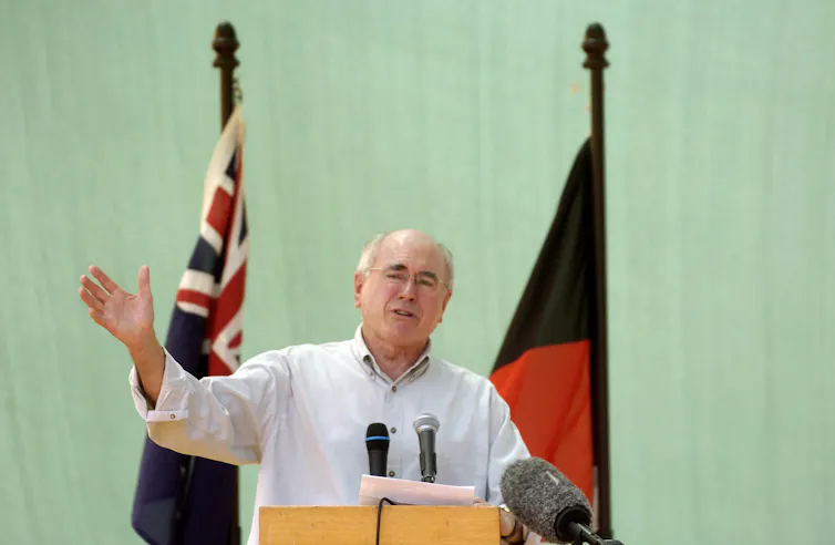 Former prime minister John Howard in a shirt with its sleeves rolled up at a podium in front of two flags