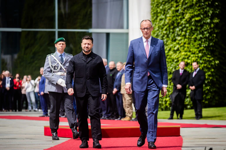 German chancellor Friedrich Merz and Ukrainian president Volodymyr Zelensky walk along a red carpet followed by a man in military uniform.