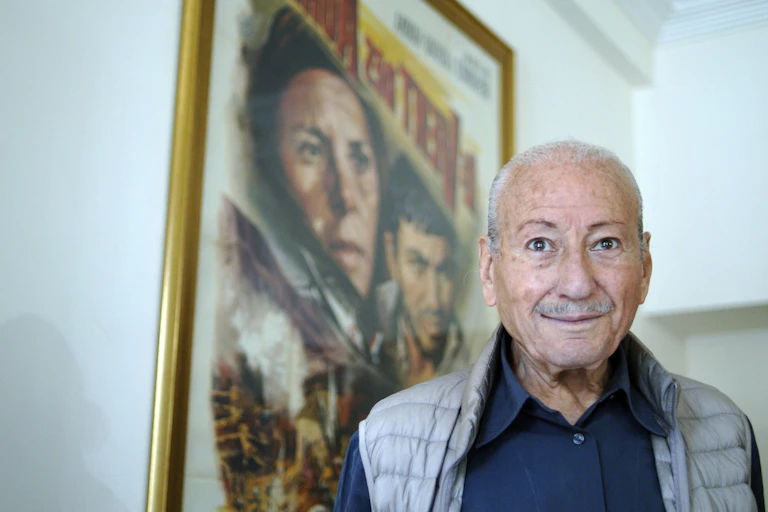 An elderly man looks mischievously to camera as he poses in front of a movie poster, hair grey.
