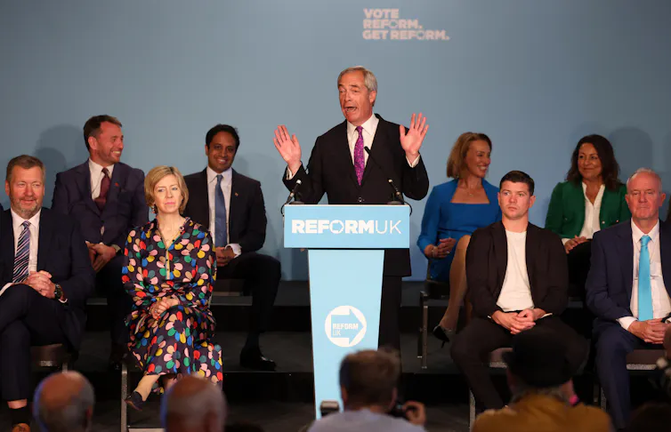 Nigel Farage speaking at a podium with Reform figures seated by him.