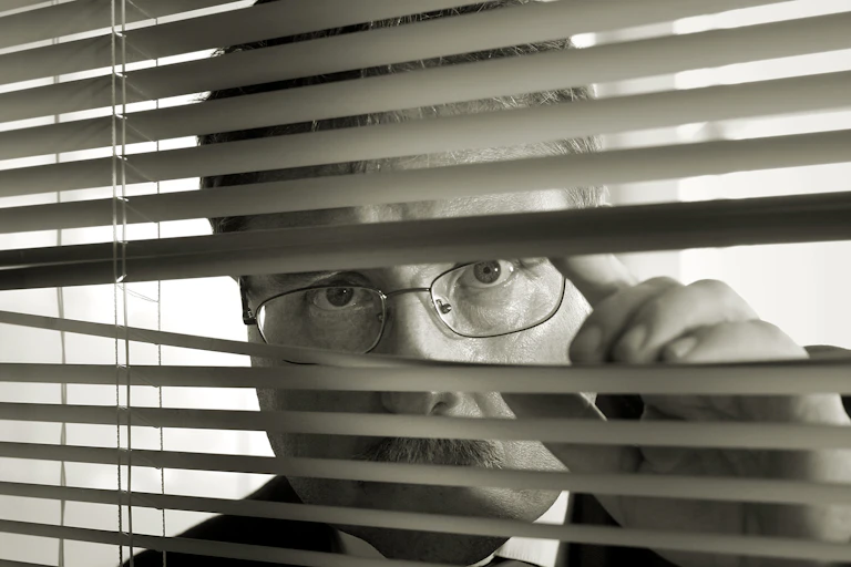 a man in a suit and glasses peers through blinds