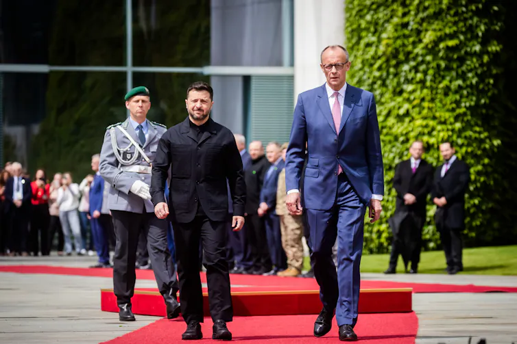 German chancellor, Friedrich Merz, walks along a red carpet with Ukrainian president, Volodymyr Zelensky with aides and military officials in the background.