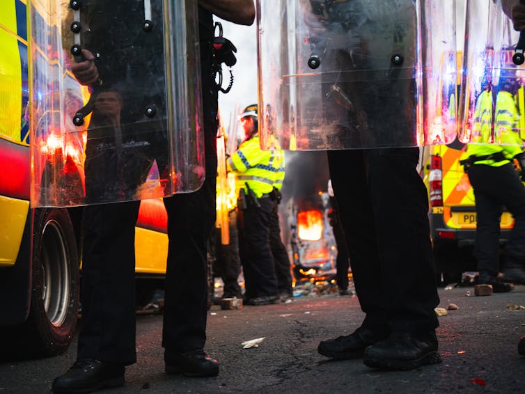 Low view of police holding riot gear, while other police and a fire burning are seen in the background