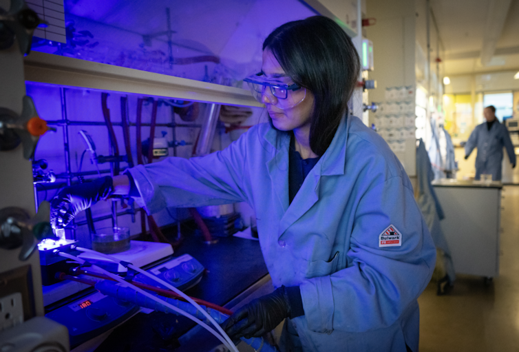 A researcher in lab goggles, gloves and a lab coat holds a bright slide over a lab bench.