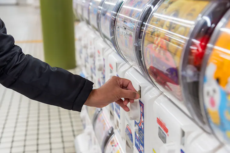 A person, seen from only the elbow down, slides a coin into the slot of a gashapon machine