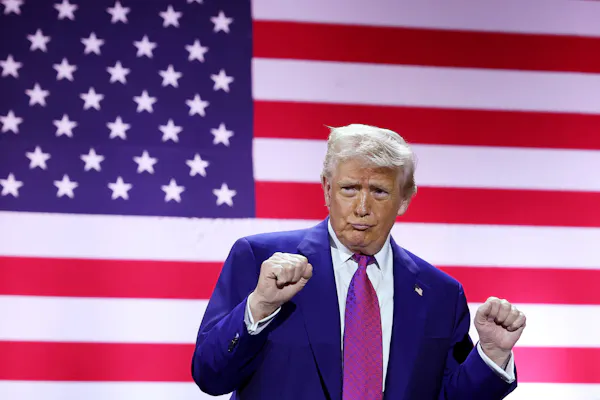 An older man with a navy blue jacket and red tie pumps his fists and stands in front of a large American flag.