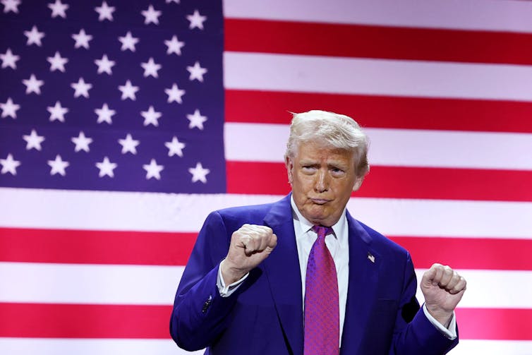 An older man with a navy blue jacket and red tie pumps his fists and stands in front of a large American flag.
