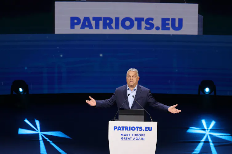 A man in a dark suit holds his arms open while standard at a podium. Behind him there is a blue backdrop that says Patriots.EU above it
