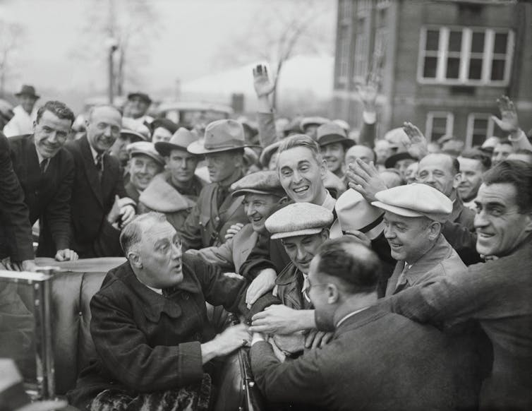 A middle-aged man in a coat and sitting in a car, is mobbed by smiling men who want to shake his hand.