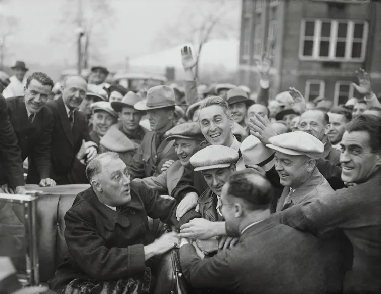 A middle-aged man in a coat and sitting in a car, is mobbed by smiling men who want to shake his hand.