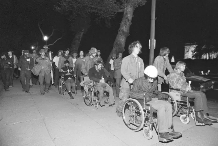 A group of men, some in wheelchairs, marching in the night along a street.