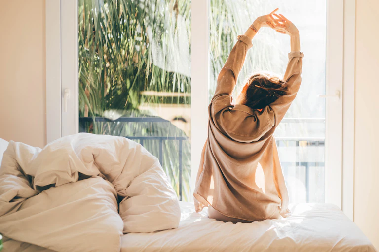 A woman stretching in bed looking out the window.