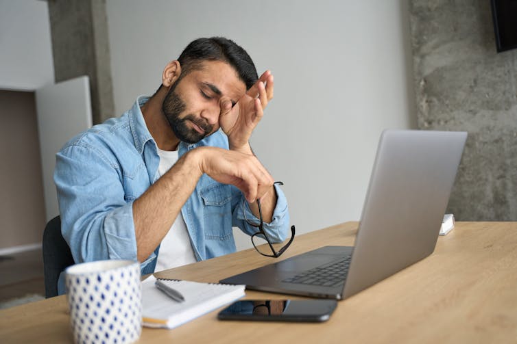A man sitting at a laptop rubbing his eye.