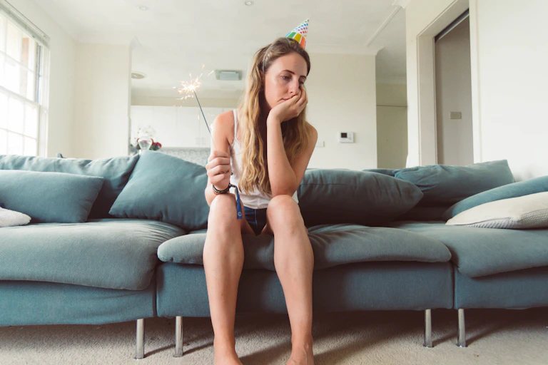 A young woman in a party hat and holding a sparkler, sits dejectedly on a couch while resting her chin in her hands