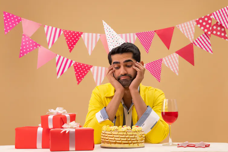 A young man surrounded by birthday decorations looks sadly at a cake in front of him