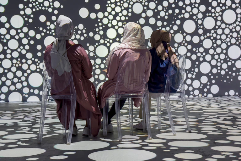Three women sit in chairs and are seen from behind looking at a digital display of white circles against a black backdrop.