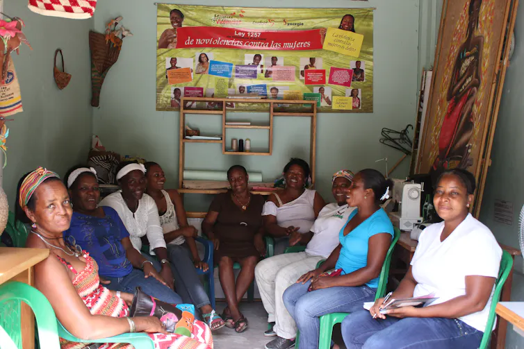A group of women smile and chat as they sit on green plastic chairs inside a small room with a large poster on one wall.
