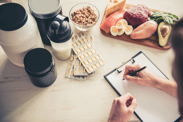 A person tracks their diet on a clipboard of paper. There is a board of meat and other health supplements on the table in front of them.
