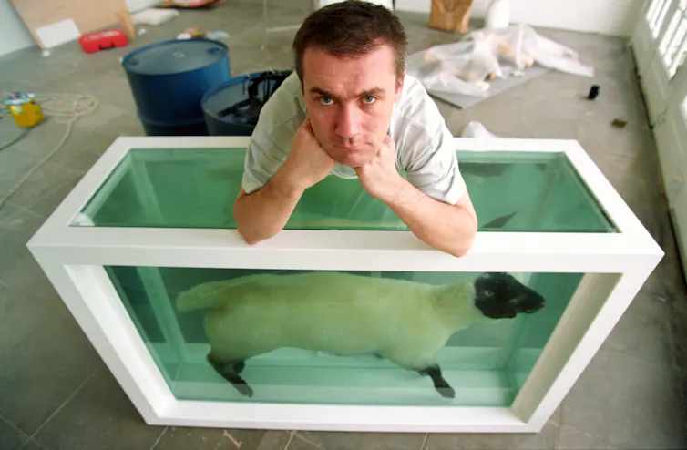 Artist Damien Hirst leaning on a tank filled with a sheep in formaldehyde.