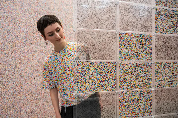 A staff member wearing a shirt with multicoloured dots blends in with Damien Hirst Currency artworks.
